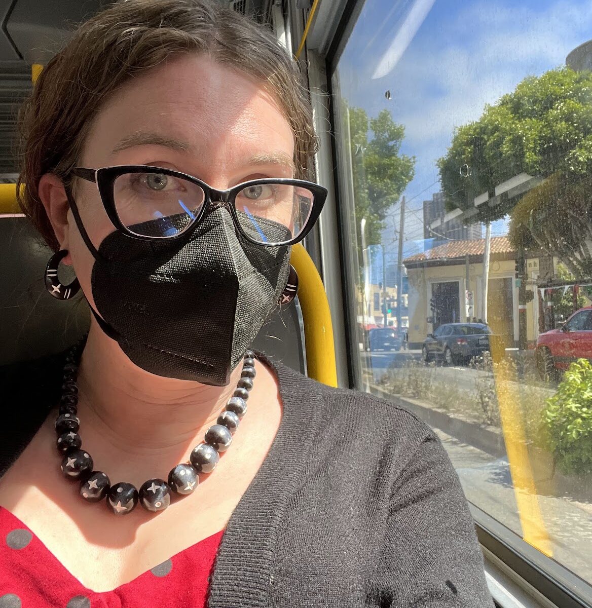 A selfie of Jen riding a bus, she's wearing a red and black polka dot top, black cardigan, and is wearing a black N95 mask.