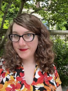 A selfie of Jen standing in front of some bushes, she has long curly brown hair and is wearing a white, black, red, and orange floral dress.